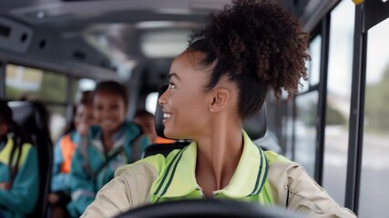 Young female driver drives bus and looks in rear view mirrors smiling. Children in school bus in background.
