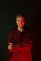 Portrait of smiling man wearing hoodie with crossed arms in neon lights against black background