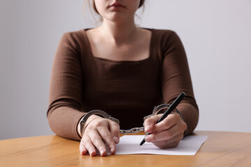 Woman in metal handcuffs writing something at wooden table against grey background, closeup
