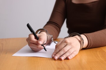 Woman in metal handcuffs writing something at wooden table against grey background, closeup