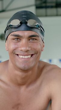 Vertical video: Male swimmer smiling following camera focus at poolside with swim cap and goggles