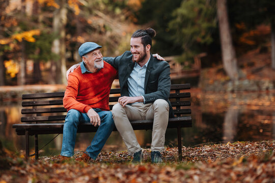 Son and older father sitting on bench by lake and talking.