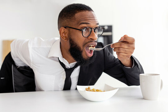 Portrait of busy African American man in glasses having breakfast in kitchen at home, wearing suit while eating cereal, rushing to office, hurry to work in the morning, being late for meeting