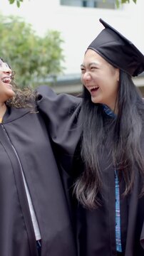 Smiling diverse teenage friends celebrating graduation outdoors on campus, wearing caps and gowns