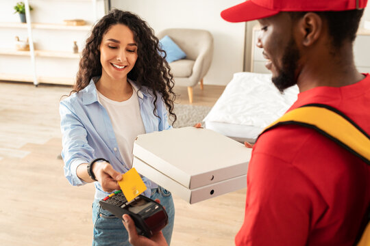 Fast Food Delivery, Digital Payment. Smiling woman using credit card, black male courier in red uniform holding POS machine in hand and giving pizza boxes to happy customer, selective focus