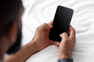Millennial african american man typing message on smartphone with blank screen, chatting on white bed in bedroom, cropped. Website and offer, ad, social distance, communication and chat, close up