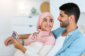 Happy islamic spouses looking at ultrasound photo of their future baby resting at home on sofa,...