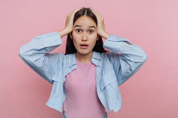 Young woman expresses shock and surprise against a pink background while holding her head with both...
