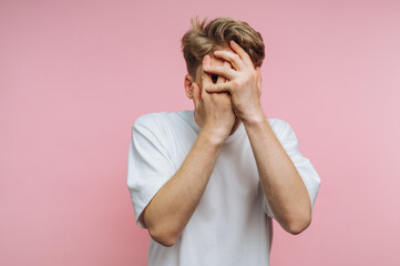 Young man expressing frustration with hands covering his face against a pink background in a...