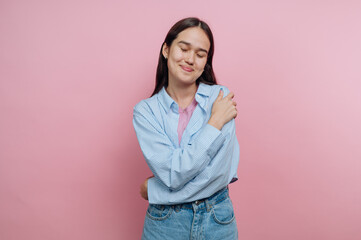 Young woman smiling softly against a pink backdrop while crossing her arms in a casual outfit,...