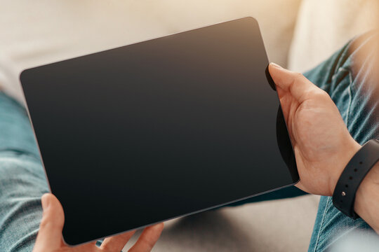 Surfing the internet. Young unrecognizable man with smartwatch holding tablet with blank screen, reading book or having video call while sitting on couch at home, cropped, closeup, mockup