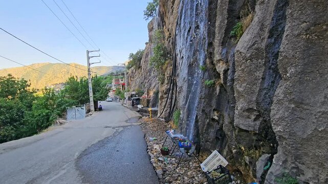 A roadside waterfall cascades down a rocky cliff under clear skies&mdash;blending natural beauty with human presence in a remote mountainous travel route.  
📍Selin village, Hawraman Takht 