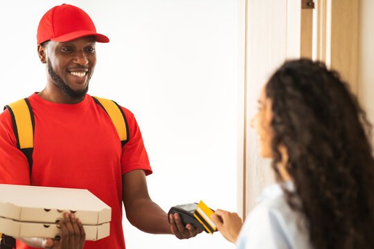 Delivery And Technology. Smiling black male courier in red uniform holding POS machine in hand and pizza boxes, woman using credit card for contactless payment, standing at door, selective focus