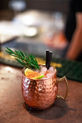 A refreshing cocktail in a beautiful copper mug, garnished with a sprig of rosemary and orange slices, set against a bar backdrop, conveying a festive or party atmosphere.
