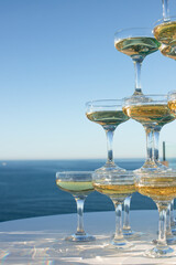 A pyramid of champagne glasses is filled with sparkling wine against a backdrop of the ocean and a clear blue sky