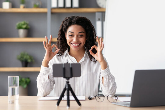 Covid-19 pandemic, education at home, excellent learning results, social distance and back to online school. Smiling young black lady teacher show ok sign with hands, look at webcam indoor interior - Powered by Adobe