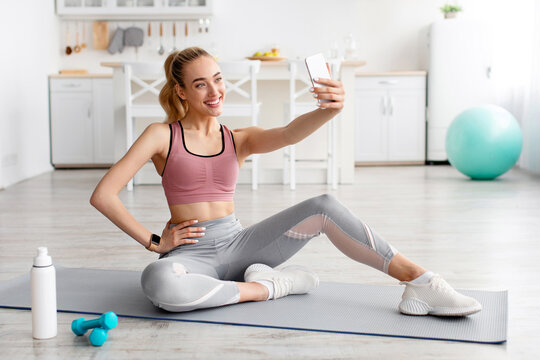 Fitness model blogger makes photo for social networks, modern device for work and workout at home. Happy young blonde lady in sportswear sits on mat takes selfie on smartphone in living room interior