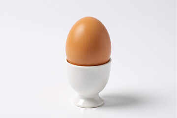 Single brown chicken egg sitting perfectly in white ceramic egg cup holder on clean white background, minimalist breakfast presentation with natural lighting and simple composition