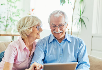 Portrait of a lovely senior mature couple together holding a tablet and having video call sitting on sofa at home