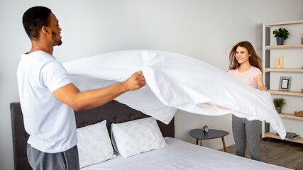 Domestic chores. Millennial multinational couple in love making bed at home, having fun while doing...