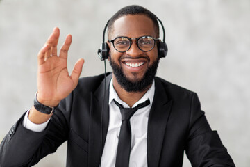 Greeting Concept. Portrait of excited young black business man in suit waving hello with hand at camera, using laptop at home office. Cheerful guy saying hi or goodbye during video call, webcam view © Prostock-studio