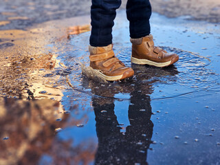 Child’s brown boots splashing in a puddle on a wet street. Close-up action shot capturing water droplets and playful movement outdoors.