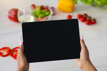 Health care, cooking at home and online call at self-isolation. Millennial african american woman hold tablet with empty screen in kitchen, at table with bright vegetables background, pov, mockup