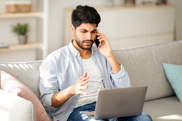 Computer problems. Displeased arab man talking on smartphone and looking on laptop screen at home, calling customer support hotline, sitting on sofa at home, free space