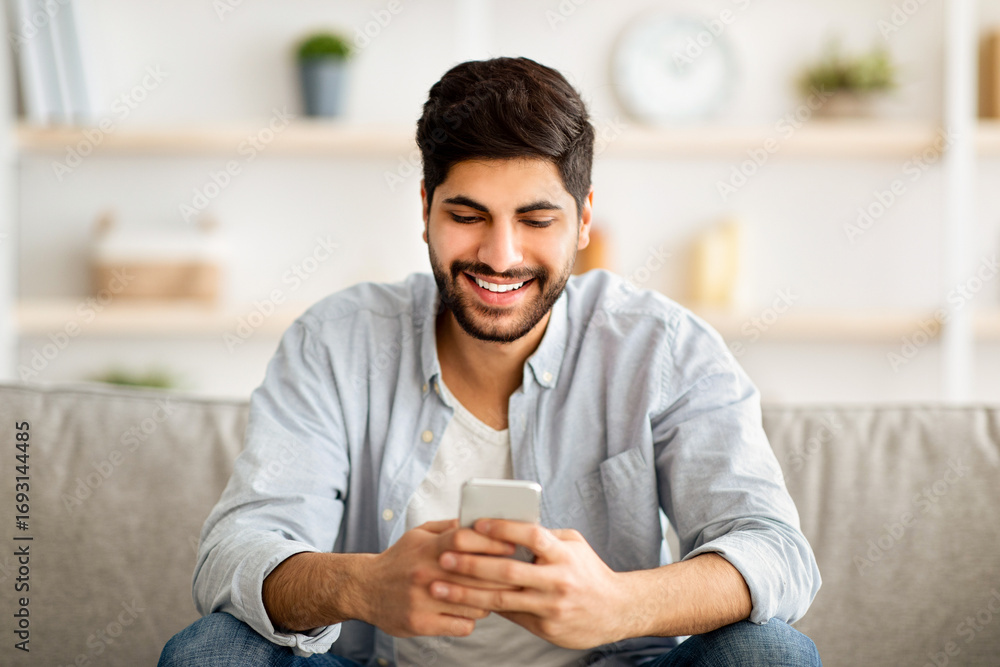 Aufkleber handsome smiling arab man sitting with smartphone in hands and messaging with friends, relaxing with