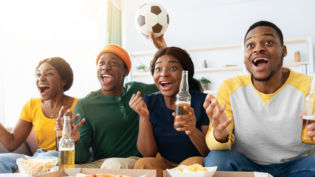 Emotional african american friends watching football match together at home, sitting on couch in living room, eating pizza and drinking beer, holding football ball, screaming and gesturing, closeup - Powered by Adobe