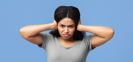 Upset african american woman closing her ears with hands, protecting from loud noise on blue studio...