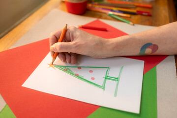 Woman&rsquo;s hand adding ornaments and star to Christmas tree drawing with colored pencils on paper.