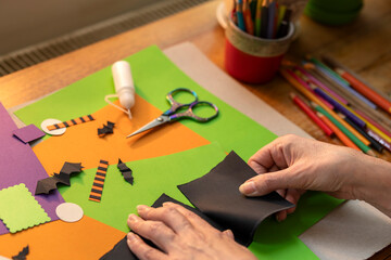 Woman’s hands folding black paper on colorful sheets with scissors, glue, and Halloween-themed...