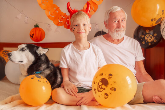 A gray-haired grandfather with his grandson and a husky dog celebrate Halloween with balloons in a decorated bedroom and smile warmly, Halloween party concept - Powered by Adobe