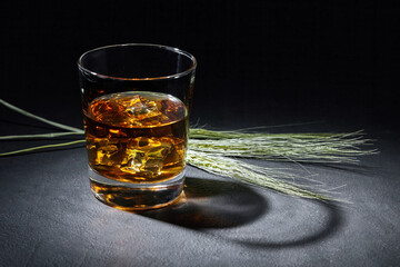 glass of amber whiskey and ears of rye on a black stone table.