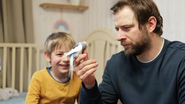 Father and son enjoying at home. Young boy and his daddy playing with funny pet budgerigar parrot. Happy family. Tamed budgie parakeet. People take care of and play with bird. Cute domestic animals