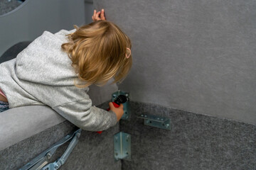 Little blonde-haired child in grey hoodie diligently focuses on assembling furniture, using...