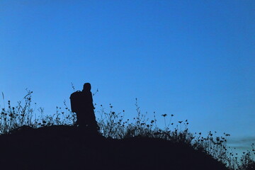 Silhouette of a lone hiker with a backpack standing on a hill at twilight, surrounded by wild grasses against a deep blue evening sky.