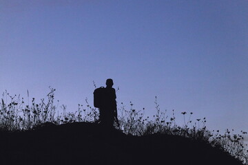 Silhouette of a lone hiker with a backpack standing on a hill at twilight, surrounded by wild grasses against a deep blue evening sky.