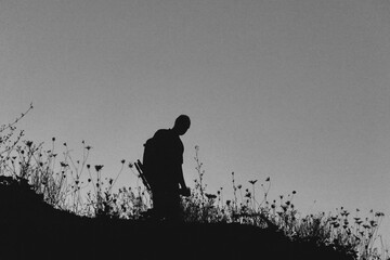 Silhouette of a lone hiker with a backpack standing on a hill at twilight, surrounded by wild grasses against a deep blue evening sky.