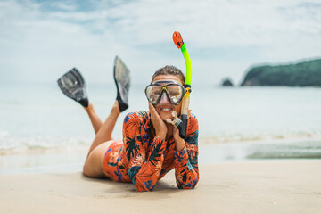 Happy female snorkeler in colorful beachwear rests on sand, holding face in hands. Enjoying coastal paradise with sea breeze and sunny weather.