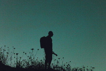 Silhouette of a lone hiker with a backpack standing on a hill at twilight, surrounded by wild grasses against a deep blue evening sky.