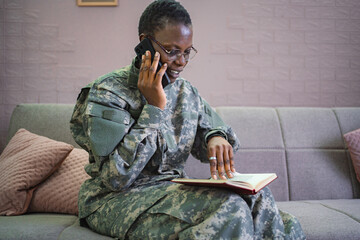 Smiling african american soldier talking on smartphone and reading book at home