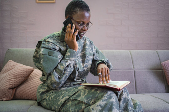 Smiling african american soldier talking on smartphone and reading book at home