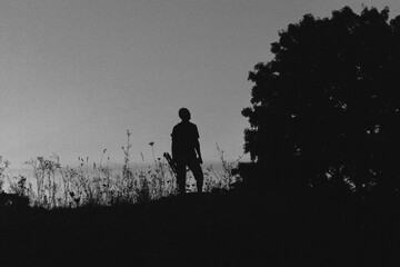 Silhouette of a lone hiker with a backpack standing on a hill at twilight, surrounded by wild grasses against a deep blue evening sky.