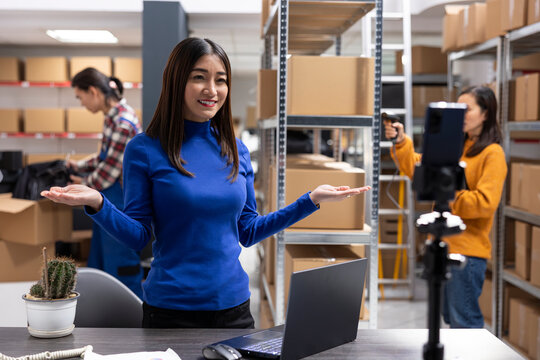 Asian employee filming a live vlog using a smartphone on a tripod, in a small business warehouse depot. Video promoting online reach, order processing and local shipping for local brand.