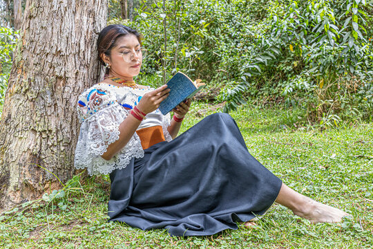 Otavalo indigenous woman barefoot, dressed in traditional embroidered attire, reading a book while resting against a tree trunk in a natural Andean setting, conveying peace and cultural connection.