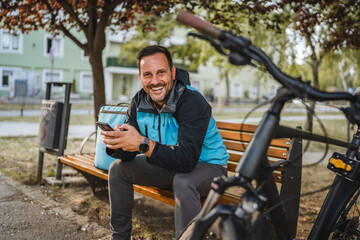 Delivery man using smart phone app sitting on bench with thermal bag and bicycle