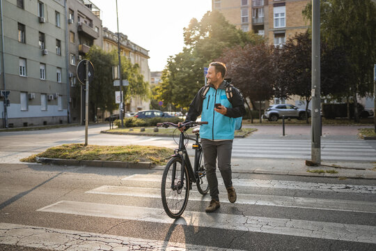 Delivery man crossing the street with bicycle and smart phone at sunset