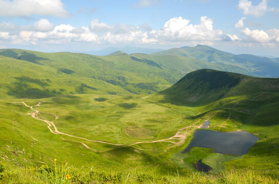Expansive view of a vast green alpine valley with a small Dogyaska lake, intersected by trails, set against a backdrop of rolling mountains and cloudy skies. Svydovets, Carpathians, Ukraine
